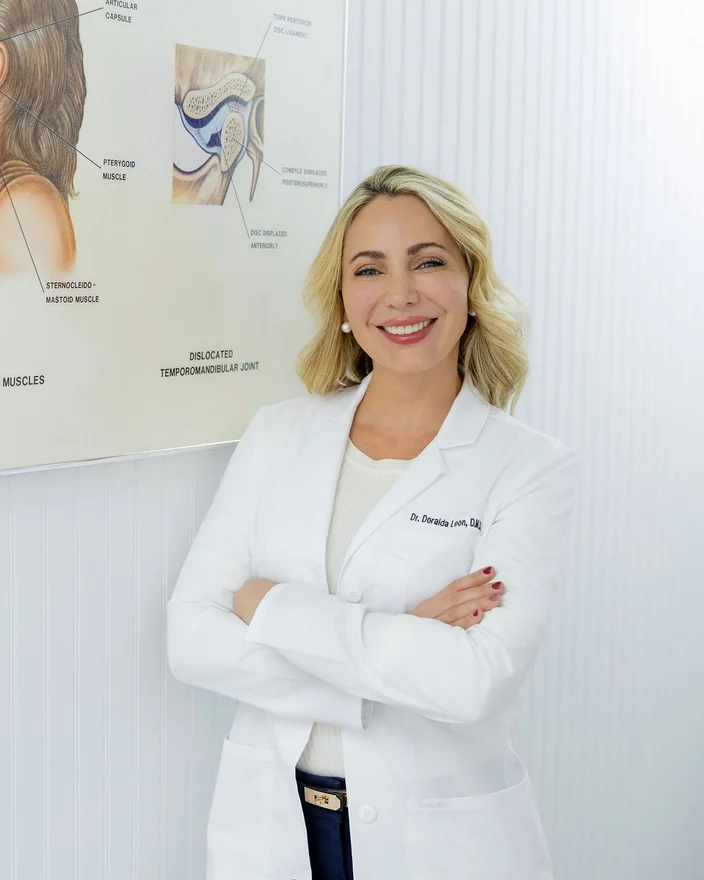 Dr. Leon, arms crossed and smiling, beside a wall chart of the jaw joint and its muscles